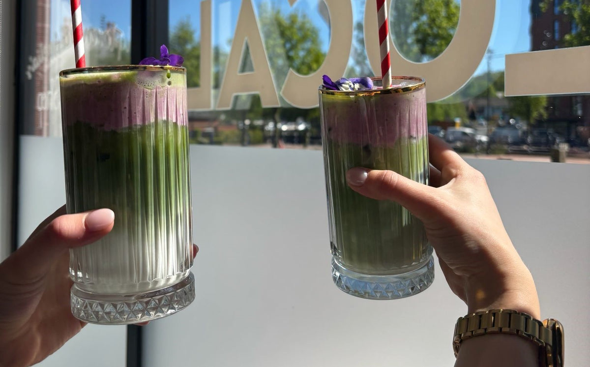 Two iced Matcha drinks with violet cold foam held up by the window