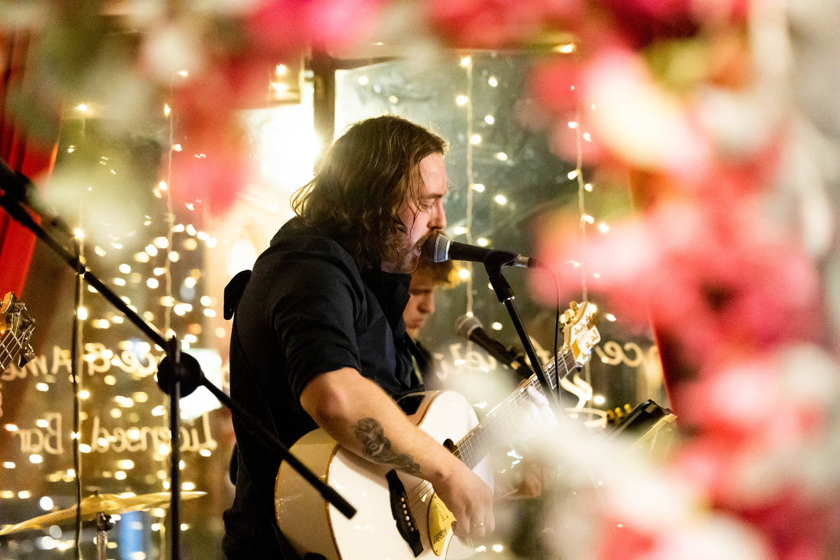 Close up of the Albert's by Florence & Amelia's in house band performing. The lead singer is playing a white guitar whilst singining into microphone. 