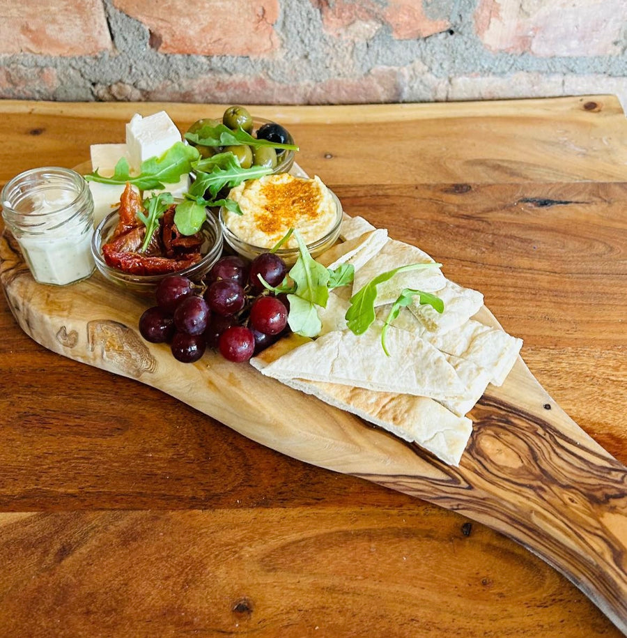 A wooden platter board with a mezze platter containing black and green olives, houmous,  toasted pitta bread, aioli, sundried tomatoes, feta cheese, chilli jam, and grapes arranged on it, placed on a wooden table.